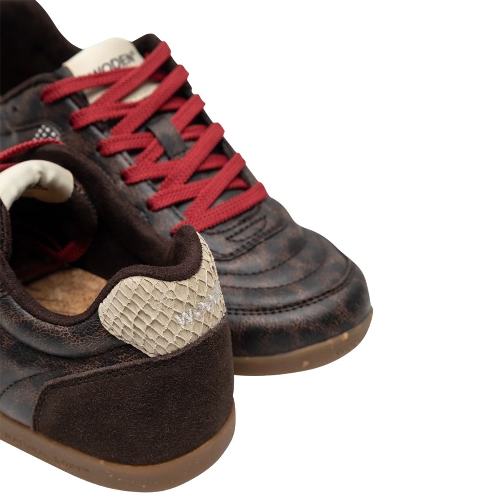 A focused view of a pair of low-top sneakers by Woden featuring dark brown textured animal print uppers, red laces, and brown gum rubber soles.