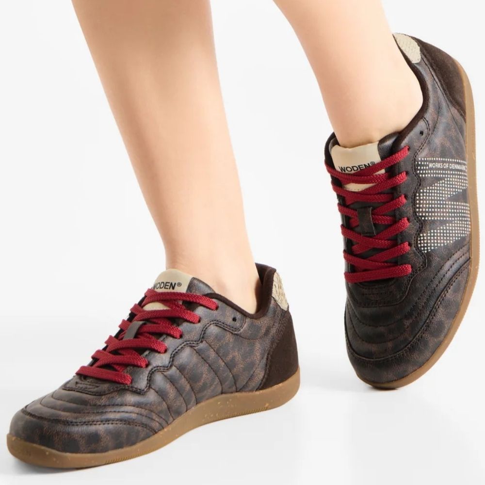 A pair of women's low-top sneakers shown on a model's feet against a clean white background. The shoes feature a dark brown and black textured leopard pattern, red laces, a beige tongue label, and a brown gum sole.