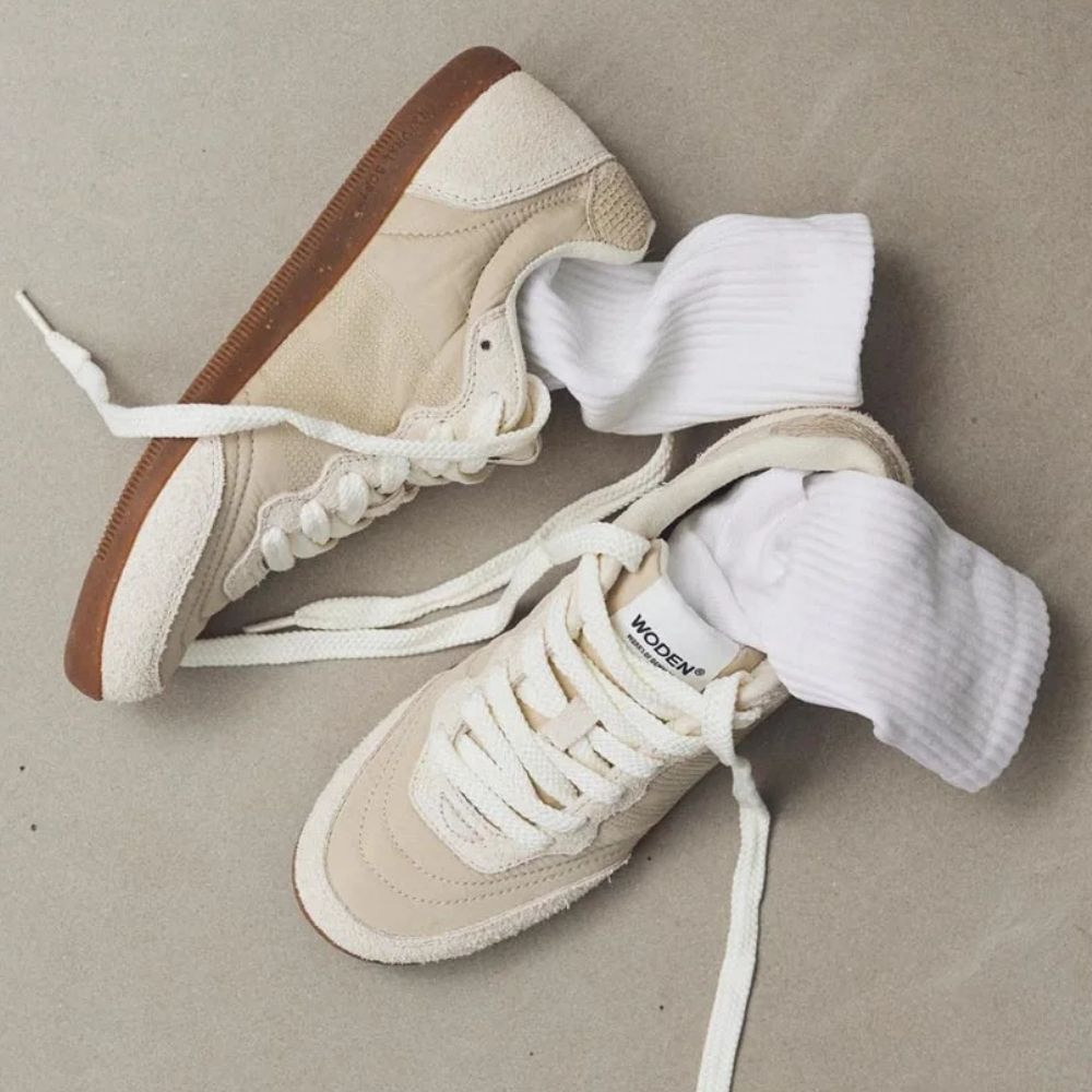 A pair of ivory low-top sneakers with white laces and brown gum soles, styled with white ribbed socks on a neutral gray background.