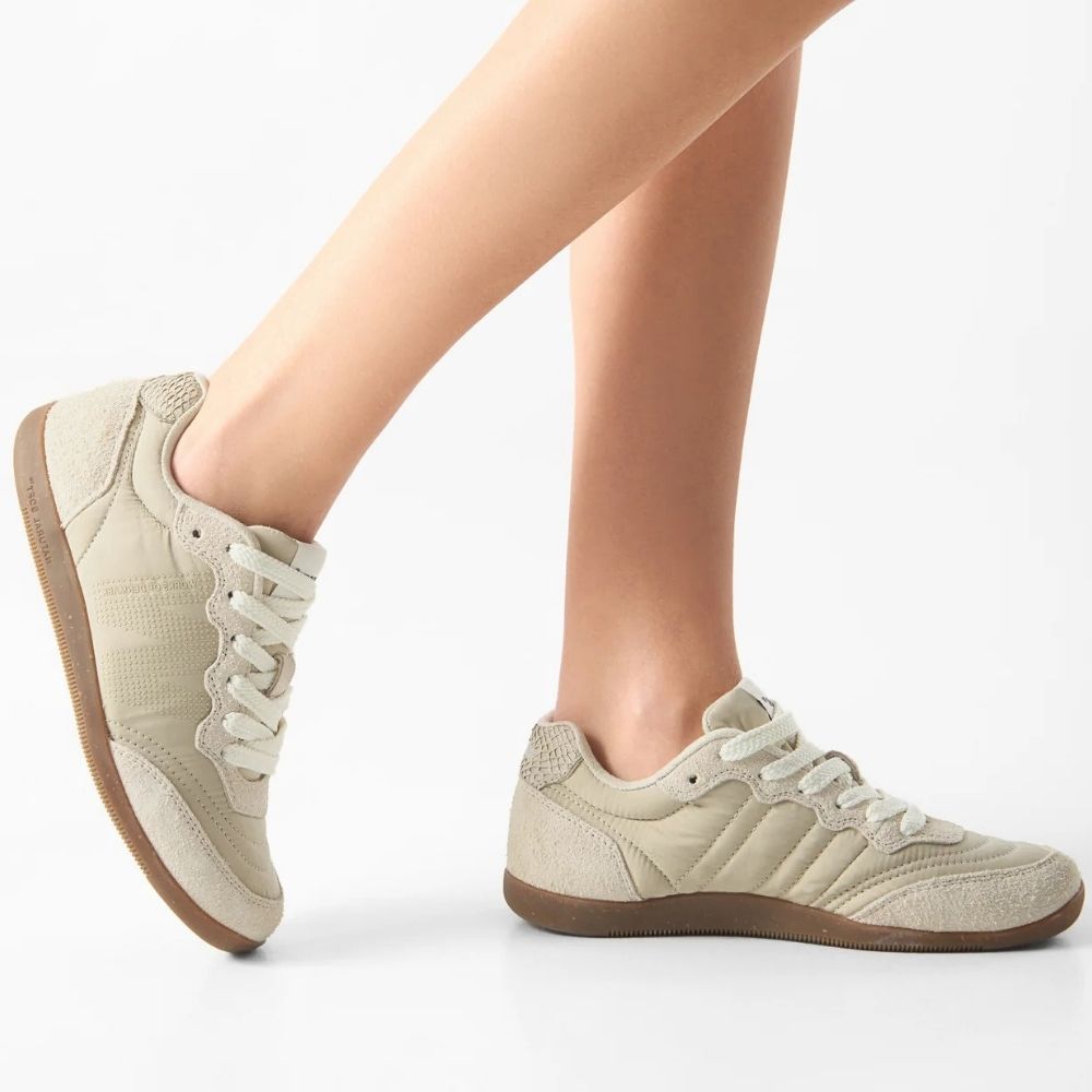 A pair of ivory low-top sneakers with textured panels and brown soles, shown on a model's feet against a clean white background.
