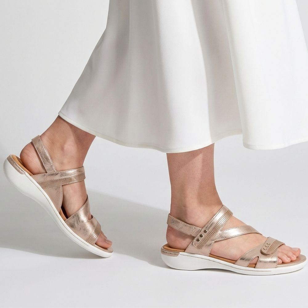 A model wearing a long white skirt and a pair of metallic rose-gold sandals with adjustable straps on a white sole, against a white studio background.