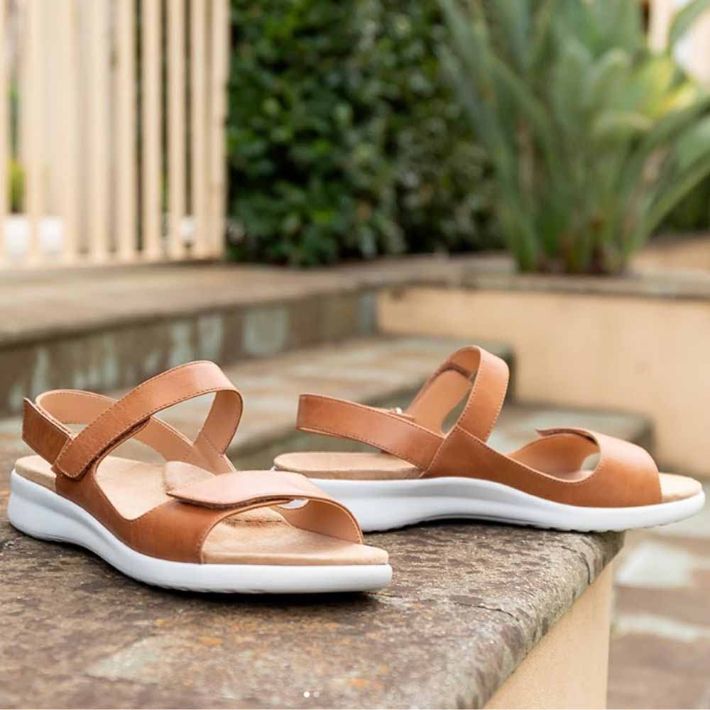 A pair of tan leather ZIERA sandals with white soles, resting on a stone ledge in an outdoor garden setting.