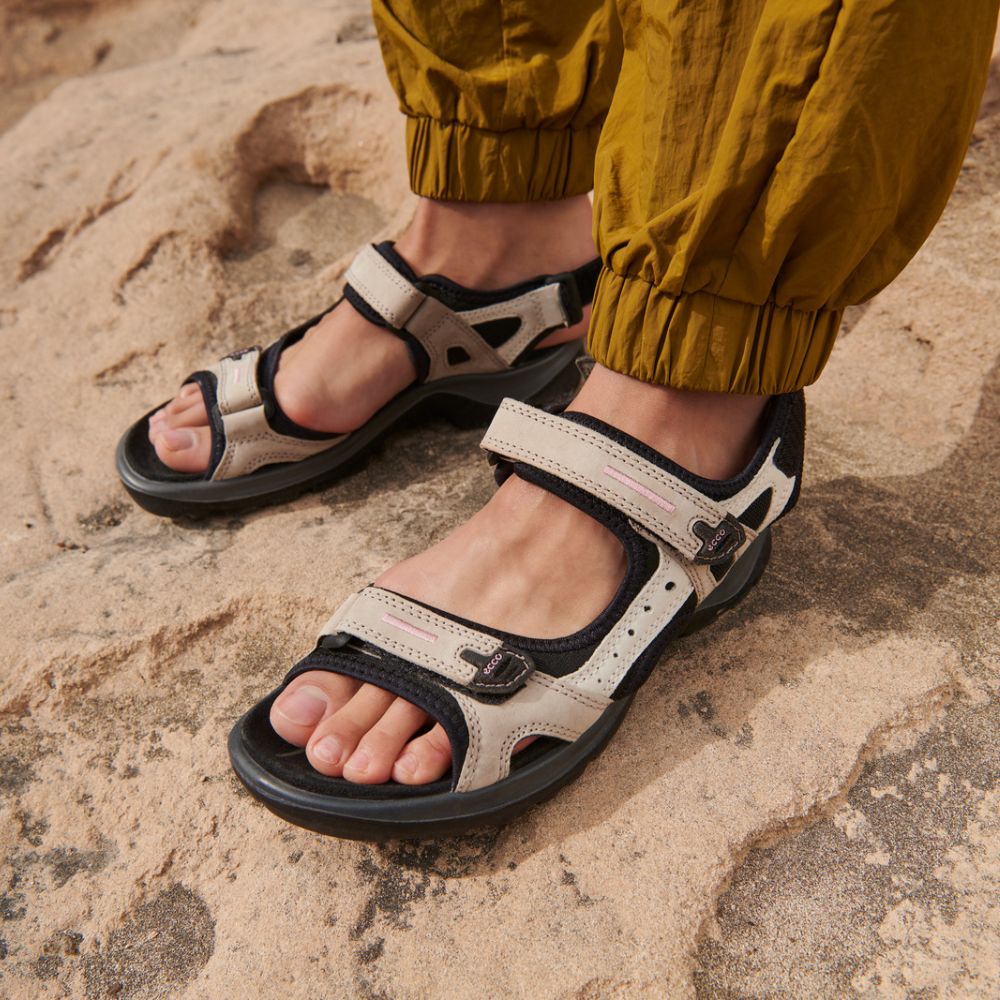 A pair of beige and black ECCO off-road sandals with Velcro straps, displayed on a rocky surface.